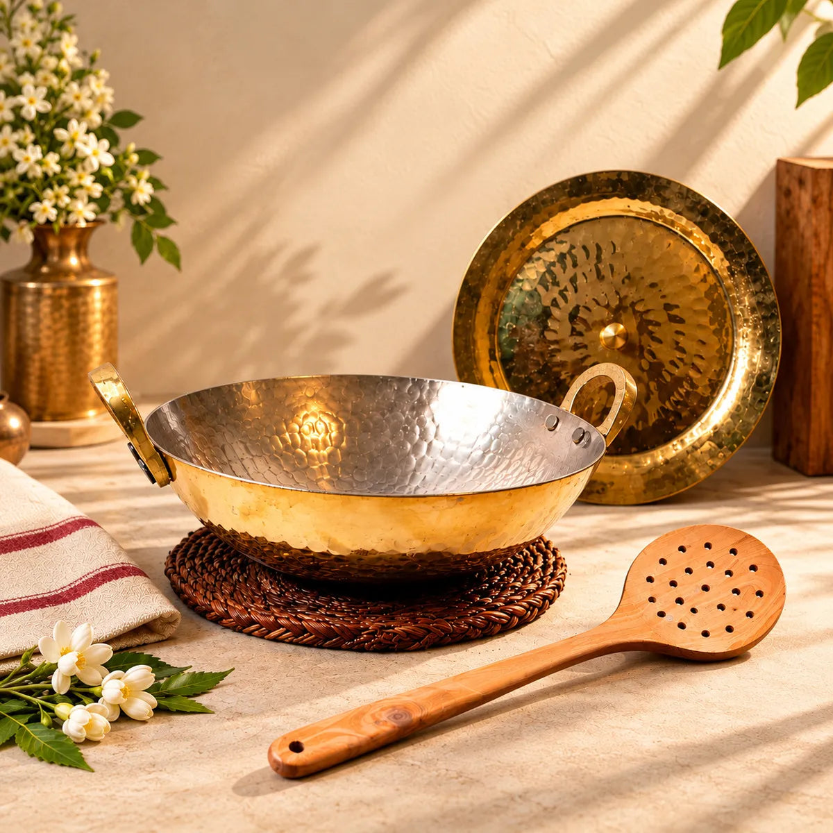 Brass kadai with lid extra large and neem wood skimmer set on a counter with jasmine flowers. Zishta Festive Cookware Combos.